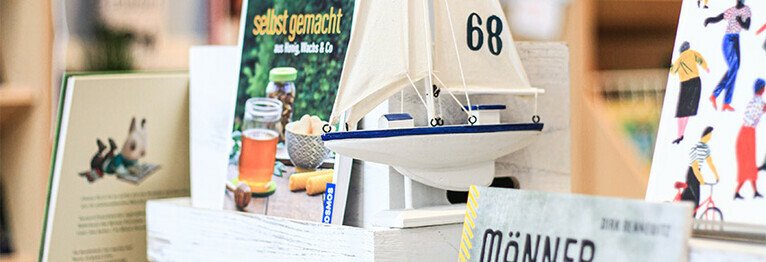 Nahansicht eines Büchertisches und einiger präsentierter Buchtitel. Ein kleines Segelschiff ist zu Dekorationszwecken auch auf dem Bild zu sehen. Die Segel sind weiß. Auf dem rechten Segel ist die Zahl 68 in dunkelbrau angebracht.