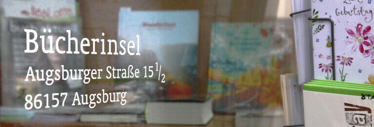 Nachaufnahme eines Teiles des Schaufensters, auf dem links im Bild der Name der Buchhandlung sowie die Anschrift angebracht sind. Also: Bücherinsel Pfersee, Augsburger Atr. 15 1/2, 86157 Augsburg. Im Hintergrund sieht man einige Bücher, die in der Auslage