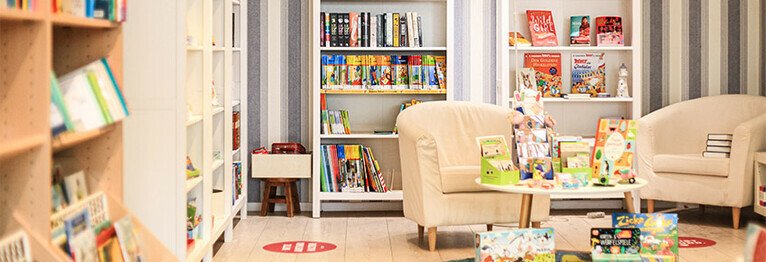 Rechts im Bild ist eine Sitzgruppe bestehend aus zwei nebeneinanderstehenden weißen Sesseln und einem kleinen, runden Couchtisch zu sehen. Auf dem Couchtisch und an den Couchtischfüßen sind Bücher zu sehen. Hinter dieser Sitzgruppe und links davon befinde