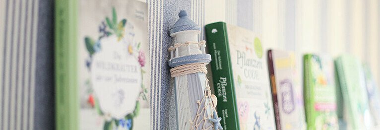 Nahansicht einer Bücherregalleiste an der blau-weiß-gestreiften Wand. Auf der Leiste stehen ein kleiner, weißer Leuchtturm, der Dekorationszwecken dient sowie einige Bücher, die zur Präsentation aufgestellt sind.